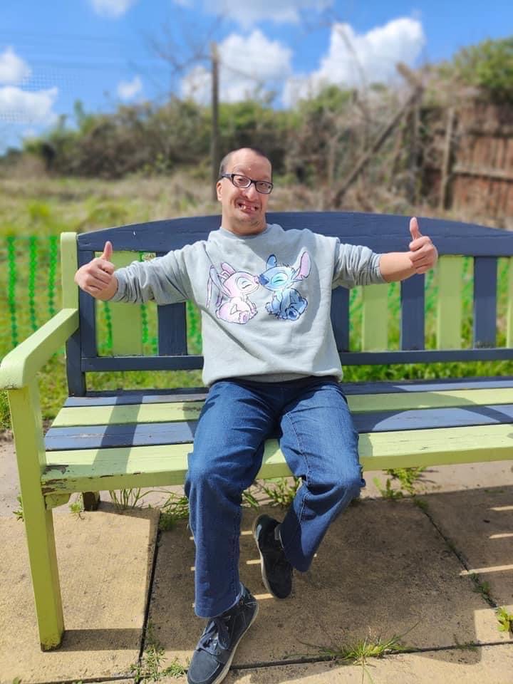 Male individual sitting outdoors on a large yellow and blue bench giving the thumbs up