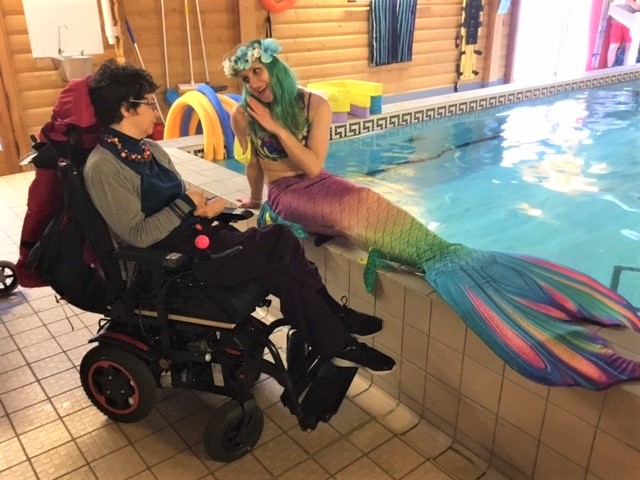 Mermaid sitting poolside beside an individual who is a wheelchair user