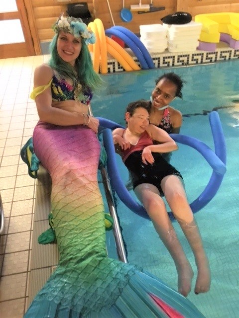 Individual being supported floating in the pool pictured with a mermaid who is sitting pool side