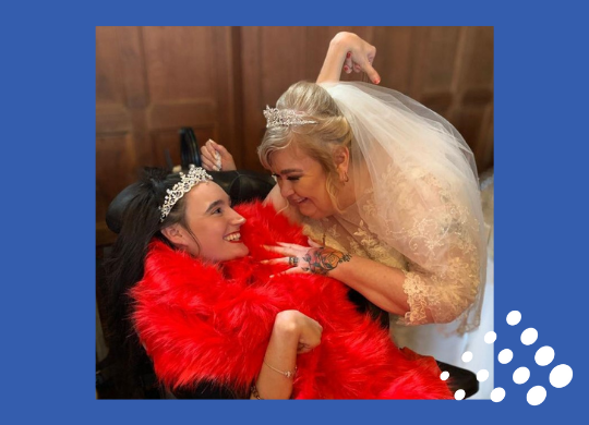 Princess Paige who is sitting in her wheelchair wearing a red dress, red stole and tiara pictured with her Mum who is wearing her wedding dress at her Mum's wedding