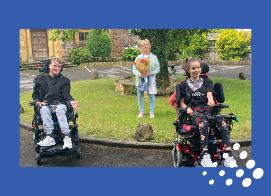 Two individuals in wheelchairs with Angela who is holding flowers standing on a lawn area celebrating 45 years at Craig Y Parc