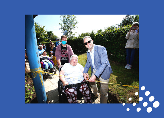 John Godden cutting the ribbon standing alongside an individual in a wheelchair with other individuals and support workers in the background at the opening of Birchwood Memorial Garden