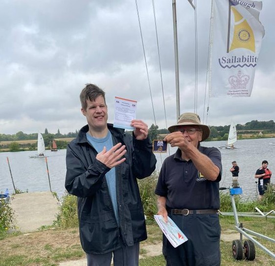 Matthew from New Road standing lakeside with his instructor smiling while holding the sailing certification he received