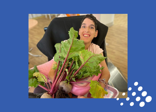 Individual sitting in a wheelchair smiling up at camera while holding large beetroots during Salutem in Bloom 2022